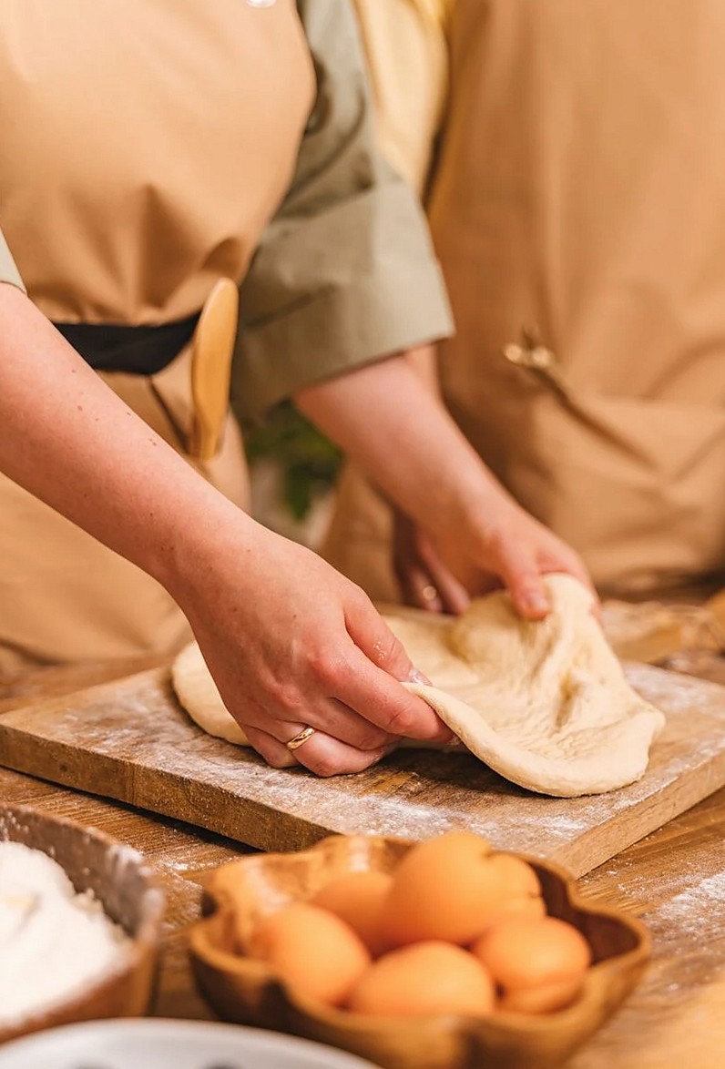 木製のボード上で生地をこねる手のクローズアップ。近くに材料が置かれ、料理をテーマにしたコンテンツに最適。
